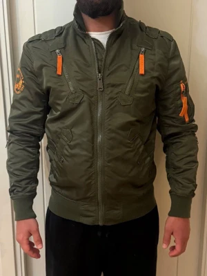 Olivgrön bomberjacka Alpha Industries - Stilren olivgrön bomberjacka från Alpha Industries med orange detaljer och broderad logga på ärmen. Jackan har ribbade muddar, dragkedjor framtill och på ärmen samt klassisk militärinspirerad look. Materialet är glansigt och slitstarkt. Priset går att diskutera 