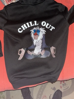 Svart hoodie med Rafiki och text - Svart hoodie med tryck av Rafiki från Lejonkungen i meditationspose och texten 'CHILL OUT' både fram och bak. Klassisk känguruficka och huva. Perfekt för dig som gillar chill vibes och Disney. Storlek s och m finns inne 