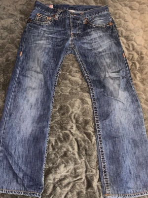 True Religion - Säljer ett par klassiska blå jeans från True Religion med kontrastsömmar och ikoniska fickdetaljer bak. Jeansen har låg midja och är tillverkade i slitstark denim med snygg tvätt. Perfekta för en avslappnad och trendig look.