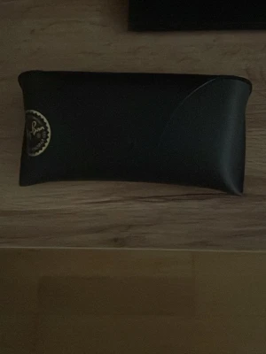 Ray-Ban svart fodral till solglasögon - Snyggt svart fodral från Ray-Ban i skinnimitation, perfekt för att skydda dina solglasögon. Klassisk design med Ray-Ban-logga i guld på framsidan. Stilren och enkel att ta med i väskan.