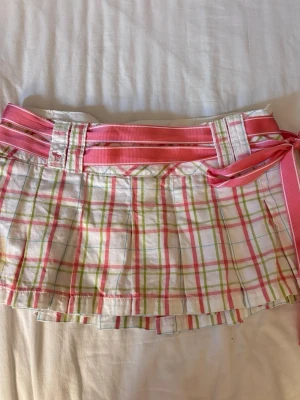 Rutig kortkjol från Abercrombie & Fitch - Superfin kortkjol i vitt med rosa, gröna och blå rutor från Abercrombie & Fitch. Kjolen har plisserad design och ett brett rosa bälte som knyts i midjan. Perfekt för dig som gillar färgglada och preppy plagg.