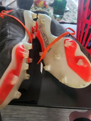 Adidas svarta fotbollsskor med orange detaljer - Säljer ett par Adidas fotbollsskor i svart med vita ränder och orange snörning. Skorna har fasta dobbar och en sula i vitt och orange. Ovandelen är i syntetmaterial och designen är smal och sportig, perfekt för snabba spelare på planen.