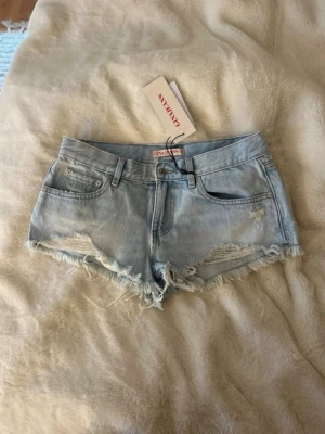 Ljusblå denimshorts med fransig kant - Säljer ett par ljusblå denimshorts med fransig, rå kant och klassisk femficksdesign. Shortsen har låg midja och stängs med knapp och dragkedja. Perfekta till sommaren för en avslappnad look.