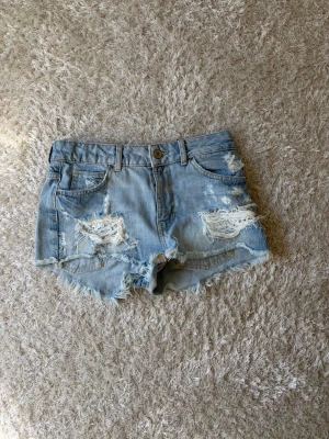 Ljusblå slitna jeansshorts - Säljer ett par ljusblå jeansshorts med slitna detaljer och fransig kant. Shortsen har klassisk femficksdesign, knapp och dragkedja framtill. 