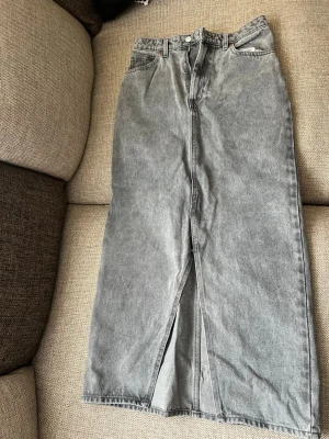 Grå jeanskjol med slits - Säljer en grå lång jeanskjol med hög midja och slits framtill. Kjolen har klassiska jeansdetaljer som bälteshällor och knappstängning. Perfekt för dig som gillar en stilren och avslappnad look. 