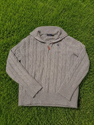 Grå kabelstickad tröja Polo Ralph Lauren - Kabelstickad grå tröja från Polo Ralph Lauren med halv dragkedja och ribbade muddar. Klassisk broderad logga på bröstet och hög krage. Perfekt för lager-på-lager och snygg till jeans eller chinos.