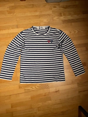 Randig långärmad tröja Comme des Garçons - Randig långärmad tröja från Comme des Garçons Play i marinblått och vitt. Tröjan har rund halsringning och två broderade hjärtan med ögon på bröstet. Perfekt för en stilren och avslappnad look. Medium men passar bättre på small (man).