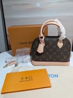 Iouis Vuitton Alma BB  - En elegant handväska i klassiskt Monogram-canvas med detaljer i ljust vachetta-läder. Väskan har den ikoniska strukturerade kupolformen och är utrustad med dubbla handtag samt en matchande clochette (nyckelhållare).