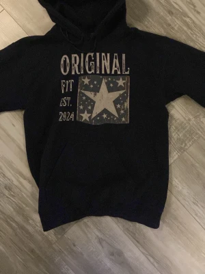 Snyg Scndi Hoodie - Svart hoodie med huva och magficka från Original Fit. Framsidan har ett stort tryck med texten 'ORIGINAL FIT EST. 2024' och en stjärna omgiven av mindre stjärnor. Perfekt för en avslappnad och cool stil.