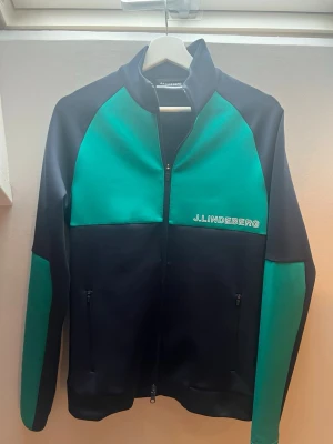J.Lindeberg ziptröja i svart och grönt -  ziptröja från J.Lindeberg i svart med gröna paneler på bröst och ärmar. Tröjan har hög krage, hel dragkedja och två fickor med dragkedja framtill. Märkeslogga på bröstet och i nacken.