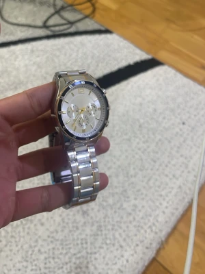 Silverfärgad kronografklocka - Stilren silverfärgad klocka med metallarmband och kronograf-funktion. Urtavlan är ljus med tre mindre visartavlor och guldiga detaljer. Perfekt accessoar för dig som gillar en klassisk och modern look.
