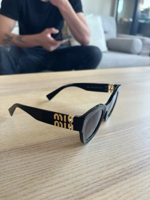 Svarta solglasögon från Miu Miu - Snygga svarta solglasögon från Miu Miu med tjock båge och guldiga detaljer på sidorna. Glasögonen har en klassisk, något kantig form och mörka glas. Perfekt accessoar för att lyfta din stil. Äkta!