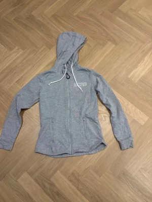 Grå hoodie från Björn Borg - Snygg grå hoodie från Björn Borg med dragkedja framtill, två fickor och vit logga på bröstet. Tröjan har huva med vita dragsnören och är tillverkad i mjukt material som känns skönt mot huden. Perfekt för en avslappnad och sportig stil.