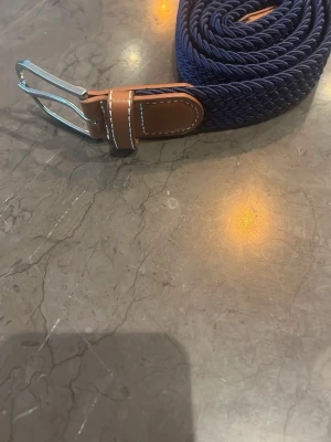 Blått flätat bälte med skinn-detaljer - Snyggt flätat bälte i mörkblått med bruna skinn-detaljer och silverfärgat spänne. Perfekt accessoar för att lyfta din outfit med en stilren touch. Passar till både jeans och chinos.