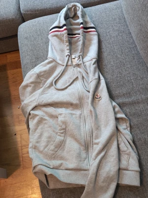 Grå zip hoodie från Moncler - Snygg grå zip hoodie från Moncler med huva och dragsko. Klassiska ränder i rött, vitt och blått på huvan samt Moncler-logga på bröstet. Mjuk bomullsblandning och två fickor framtill. Perfekt för en avslappnad och trendig look.