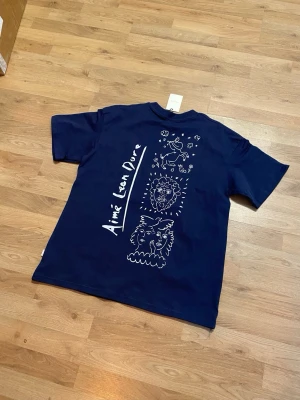 Mörkblå Aimé Leon Dore t-shirt - Snygg mörkblå t-shirt från Aimé Leon Dore med unikt vitt grafiskt tryck på ryggen som visar ansikten, ett lejon och en häst. T-shirten har normal passform och korta ärmar. Perfekt för dig som gillar streetwear och coola prints.