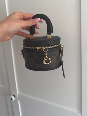 Coach mini handväska i svart och brunt - Superfin mini handväska från Coach i svart och brunt konstläder med guldiga detaljer och dragkedja. Väskan har rund form, ett litet handtag och Coach-logga framtill. Perfekt för dig som gillar trendiga accessoarer och vill sticka ut med en unik modell. KÖPT UTOMLANDS