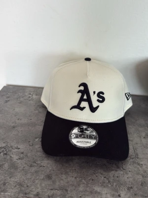 New Era 9FORTY A's keps svart/vit - Snygg 9FORTY keps från New Era med Oakland A's logga framtill. Kepsen är vit med svart skärm och justerbar passform. Klassisk basebollstil med broderad logotyp och bekvämt material. Perfekt för dig som gillar sportig streetwear. Aldrig använt!