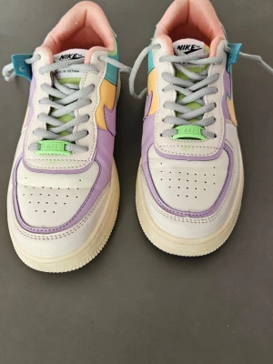 Nike Air Force 1 Pastell Sneakers -  Nike Air Force 1 Shadow "Pale Ivory" Klassisk sneaker i en lekfull design med dubbla lager, detaljer i pastellfärger (bland annat ljuslila, gult, ljusblått och mintgrönt) samt en något tjockare mellansula än originalet. Modellen släpptes 2019 och är numera slutsåld i butik. Storlek: 39 Skick: Sparsamt använda Material: Ovandel i läder och syntet, foder i textil och sula i gummi  Stilkod: CI0919-101  Övrigt: Säljs utan originalkartong