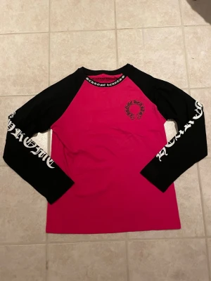 Chrome Hearts Longsleeve - Säljer en långärmad t-shirt från Chrome Hearts med svart tryck på ärmarna och logga på bröstet. Tröjan har svart halsringning och ärmar, samt en stark rosa kropp. Perfekt för dig som gillar streetwear och vill sticka ut.