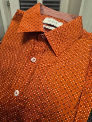 Orange mönstrad skjorta från Jack's comfort fit M - Snygg orange skjorta från Jack's med små geometriska mönster i vitt och svart. Skjortan har klassisk krage, vita knappar och lång ärm. Perfekt för dig som vill sticka ut med färg och mönster. Tillverkad i bomull för en skön känsla.