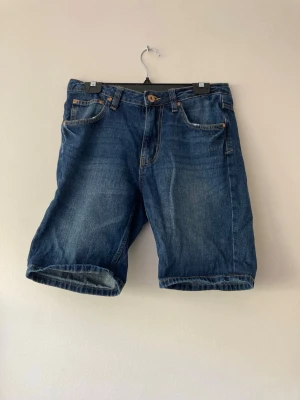 Blå jeansshorts i klassisk modell - Säljer ett par blå jeansshorts i klassisk femficksmodell med normal passform. Shortsen har knapp och dragkedja framtill samt bälteshällor. Perfekta för sommaren och enkla att matcha med olika stilar.