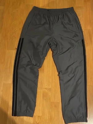 Grå Adidas mjukisbyxor med svarta ränder - Snygga grå mjukisbyxor från Adidas med klassiska svarta ränder längs sidorna. Byxorna har resår i midjan och vid benslut, samt diskret Adidas-logga på ena benet. Perfekta för chill eller träning.