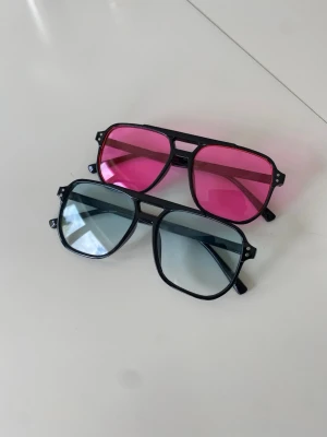 Två par retro solglasögon svart båge - Två par solglasögon med svart ram och stora fyrkantiga glas. Ena paret har rosa tonade glas och det andra har gråtonade glas. Snygg retrostil med breda skalmar, perfekta för att sticka ut i mängden.