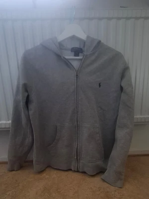 Grå hoodie från Polo Ralph Lauren - Klassisk grå hoodie med dragkedja från Polo Ralph Lauren. Tröjan har huva, två fickor framtill och den ikoniska lilla broderade loggan på bröstet. Perfekt för en avslappnad och stilren look. Mjuk bomullsblandning för extra komfort.