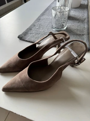 Pumps - Säljer ett par bruna pumps från berskha med spetsig tå och öppen häl. Skorna har en elegant slingback-design med justerbar rem och är tillverkade i mockaliknande material. Perfekt för dig som gillar stilrena och trendiga skor. Helt nya men säljer då jag råkade ta fel storlek!💕