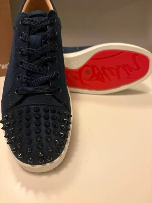 Mörkblå Louboutin sneakers med nitar - Snygga mörkblå sneakers från Christian Louboutin i mocka med svarta nitar på tån och klassisk röd sula. Skorna har vita sulor och mörkblå snören, perfekt för dig som vill sticka ut med en edgy och lyxig stil. Dock är en spik av, och har lite ferg på de vita delen men är lätt att fixas! 