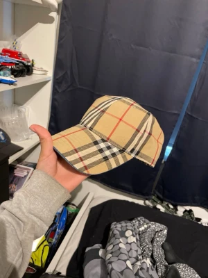 Beige keps med rutigt mönster Burberry - Snygg beige keps från Burberry med klassiskt rutigt mönster i svart, vitt och rött. Kepsen har böjd skärm och justerbar baksida. Perfekt accessoar för en trendig och stilren look.