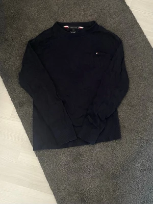 Mörkblå stickad tröja Tommy Hilfiger - Snygg mörkblå stickad tröja från Tommy Hilfiger i pima bomullsblandning med cashmere. Klassisk modell med rund hals och liten broderad logga på bröstet. Perfekt för dig som gillar stilrena och bekväma plagg.
