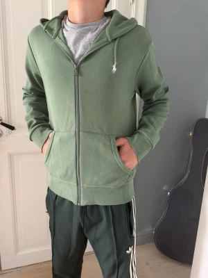 Grön hoodie från Polo Ralph Lauren - Snygg grön hoodie från Polo Ralph Lauren med dragkedja framtill och klassisk huva. Tröjan har två fickor på magen och den ikoniska vita Polo-loggan broderad på bröstet. Tillverkad i mjuk bomullsblandning som känns skön mot huden. Perfekt för en avslappnad stil.