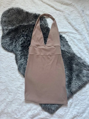 Beige halterneck kortklänning - Trendig beige kortklänning med djup v-ringning och halterneck. Klänningen har en kroppsnära passform och är tillverkad i ett mjukt, ribbat material som ger en snygg siluett. Perfekt för dig som gillar minimalistisk och stilren design.