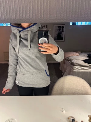 Grå hoodie från ION med hög krage - Säljer en snygg grå hoodie från ION med hög krage och dragsko. Hoodien har blå detaljer vid fickan och insidan av kragen, samt en liten logotyp nertill. Perfekt för en chill och sportig stil.