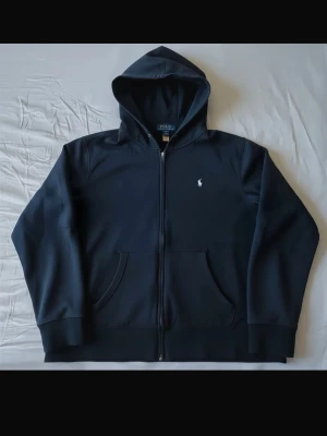 Marinblå hoodie från Polo Ralph Lauren - Snygg marinblå hoodie med dragkedja från Polo Ralph Lauren. Klassisk design med huva, fickor framtill och den ikoniska vita logon broderad på bröstet. Tillverkad i mjuk bomull för skön komfort och stilren look.