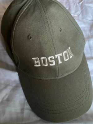 Olivgrön keps med BOSTON-brodyr - Snygg olivgrön keps med vit brodyr där det står 'BOSTON' framtill. Klassisk böjd skärm och justerbar rem baktill. Tillverkad i bomullstyg som ger en avslappnad och cool look.