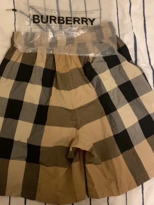 Burberry rutiga shorts med snörning - Snygga Burberry shorts med klassiskt rutigt mönster i beige, svart, vitt och rött. Shortsen har elastisk midja med svart snörning och är tillverkade i ett lätt bomullsmaterial. Perfekta för en trendig och avslappnad stil.