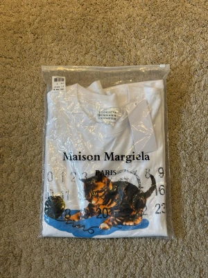 Maison margiela vit t-shirt med kattmotiv - Maison Margiela t-shirt i vitt med tryck av en lekande katt och garnnystan framtill. T-shirten har rund hals och korta ärmar. Märkets logga och siffror är tryckta på framsidan. Perfekt för dig som gillar unika designer.