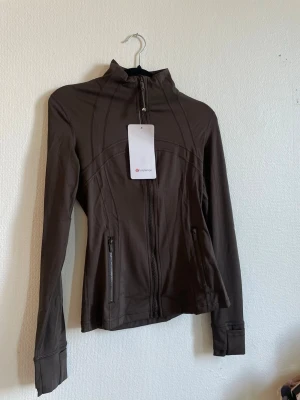 Brun jacka från Lululemon st 2  - Snygg brun jacka från Lululemon med hel dragkedja framtill och två fickor med dragkedja. Jackan har lång ärm och en figurnära passform, perfekt för en sportig och stilren look.