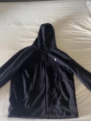 Svart hoodie från Polo Ralph Lauren - Svart hoodie med dragkedja från Polo Ralph Lauren. Klassisk design med vit broderad logga på bröstet, huva med dragsko och två fickor framtill. Perfekt för en avslappnad och stilren look.