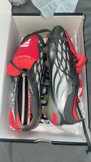 Adidas Predator fotbollsskor grå/röd - Snygga Adidas Predator fotbollsskor i grå, vitt och rött med klassisk design och tydliga detaljer. Skorna har mönstrad ovandel, röda plösar med Adidas-logga och text, samt robusta dobbar för bra grepp på planen. Perfekta för dig som vill sticka ut på matchen.