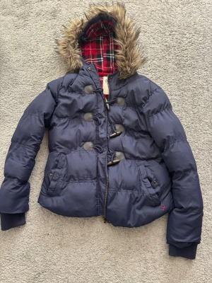 Mörkblå pufferjacka med fuskpäls - Snygg mörkblå pufferjacka från Denim Co. med huva kantad av fuskpäls och röd- och svartmönstrat rutigt foder. Jackan har dragkedja, knäppning med träknappar och två fickor framtill. Perfekt för kalla vinterdagar.