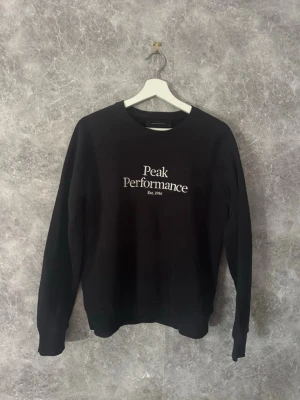  sweatshirt Peak Performance - Snygg svart sweatshirt från Peak Performance med vit logotyp och text på bröstet. Tröjan har rund halsringning, långa ärmar och ribbade muddar vid ärmslut och nederkant. Perfekt för en avslappnad och sportig stil.