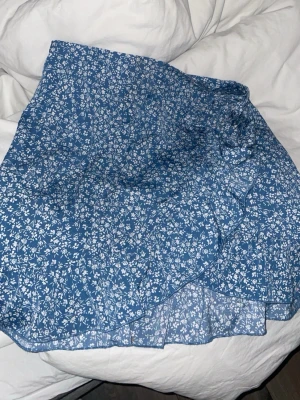 Blå blommig omlottkjol - Söt blå kortkjol med vitt blommönster och omlottdetalj. Kjolen har volangkant och är perfekt för varma dagar. Lätt och luftig känsla, passar till både sneakers och sandaler.