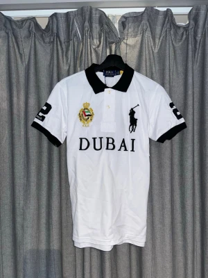 Vit pikétröja Polo Ralph Lauren Dubai - Vit pikétröja från Polo Ralph Lauren med svart krage och svarta detaljer. Stort tryck med texten DUBAI och broderad logga på bröstet, samt siffran 2 på ärmar och rygg. Klassisk polokrage och korta ärmar.