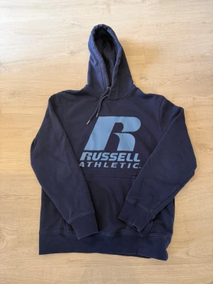 Mörkblå hoodie från Russell Athletic - Snygg mörkblå hoodie med stor ljusblå Russell Athletic-logga på bröstet. Klassisk huva med dragsko och ribbade muddar vid ärmslut och nederkant. Perfekt för en avslappnad och sportig stil.