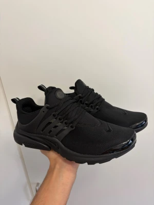 Nike Presto Triple Black sneakers - Svarta Nike Presto sneakers med mesh-ovandel och plastdetaljer vid snörningen. Skorna har en chunky sula och diskret swoosh-logga på sidan och tån. Lätta och flexibla med sportig vibe, perfekta för en clean streetstyle-look.