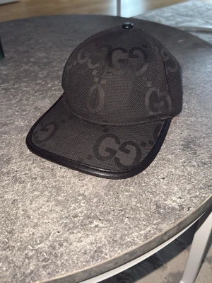 Svart Gucci keps - Snygg svart keps från Gucci med klassiskt GG-mönster över hela kepsen. Kepsen har böjd skärm med läderdetalj längs kanten och är tillverkad i ett slitstarkt tygmaterial. Perfekt för dig som vill ha en stilren accessoar med lyxig känsla.